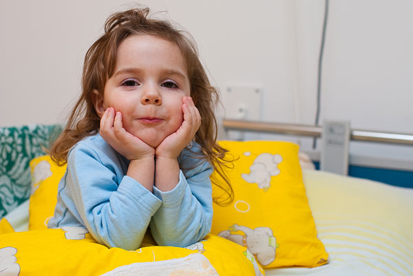 Ein kleines Mädchen mit braunen Haaren sitzt auf einem Bett, stützt den Kopf auf die Fäuste und schaut lächelnd in die Kamera. Um sie herum sind gelbe Kissen.